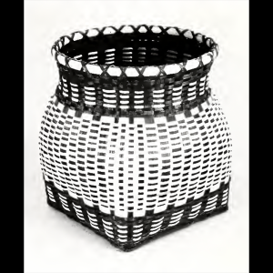 Black and white photo of a Cherokee basket. The basket has a square bottom that curves like a vase into a circular opening at the top. It uses mostly light colored material with dark material used to create patterned designs in the weave along the bottom and top of the basket.