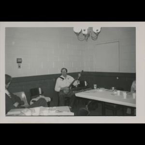 A black and white photo with a border shows Clark Jones seated by a fold out table. He is holding a banjo with a white button up, glasses, and a shoulder strap for the instrument. Various food plates and cups are on the table and one other person can be seen in the corner of the photo watching Clark Jones