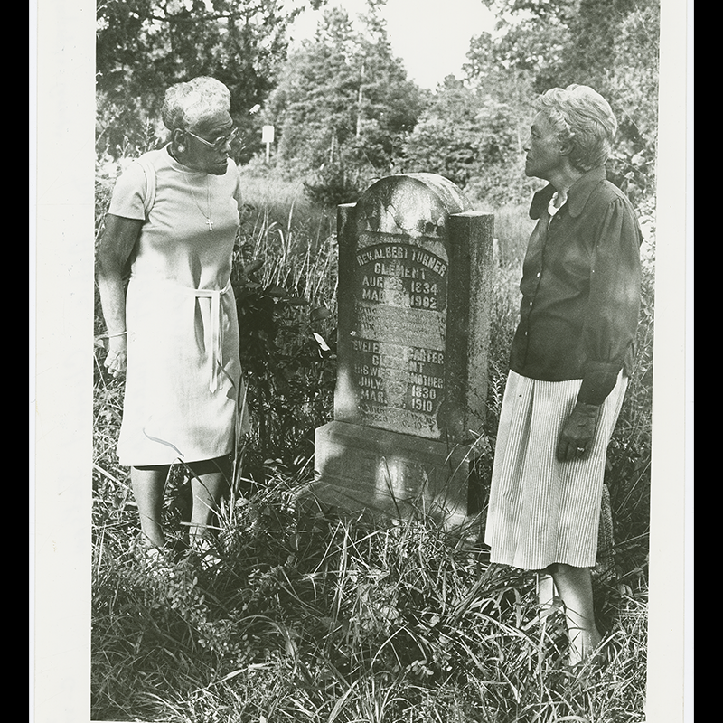 Reverend Albert Clement’s granddaughters, Dr. Abbie Clement Jackson and Ruth Clement Bond, stand on either side of his gravesite in the St. John's AME Zion Church cemetery. This tombstone shows a dove and ornamental design around it, as well as a script that covers the rest of the tombstone. It can clearly read his name, birth date, and death date, as well as that for his wife. There are carvings that appear to be script on the rest of the tombstone, but it is difficult to determine what it may say.