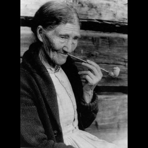 Sophie Campbell shown in a black and white photo. She is an elderly woman with her hair pulled back and wearing a knit jacket. She is holding a clay pipe in one hand and posing for the photo as if she was taking a puff. The background is a wall with wooden planks.