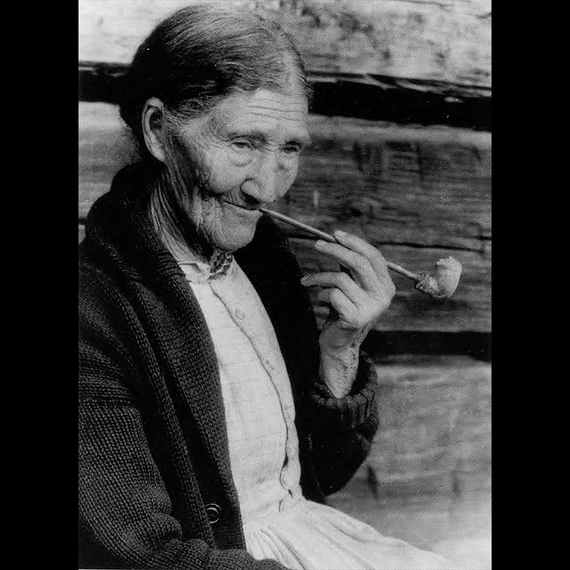 Sophie Campbell shown in a black and white photo. She is an elderly woman with her hair pulled back and wearing a knit jacket. She is holding a clay pipe in one hand and posing for the photo as if she was taking a puff. The background is a wall with wooden planks.