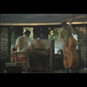 Yusuf Salim is seen wearing a shirt with traditional African-style print, his back is to the camera as he plays at an upright wooden piano. Beside him facing the camera is a person playing a bass. They are on a stage in the park.