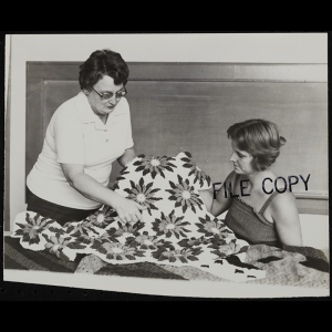 A black and white photo with the words “File Copy” stamped on the right side of the photo. Behind a table one person is seated and the other stands while they hold a blanket with a star pattern and are posed for a candid photo in the classroom.