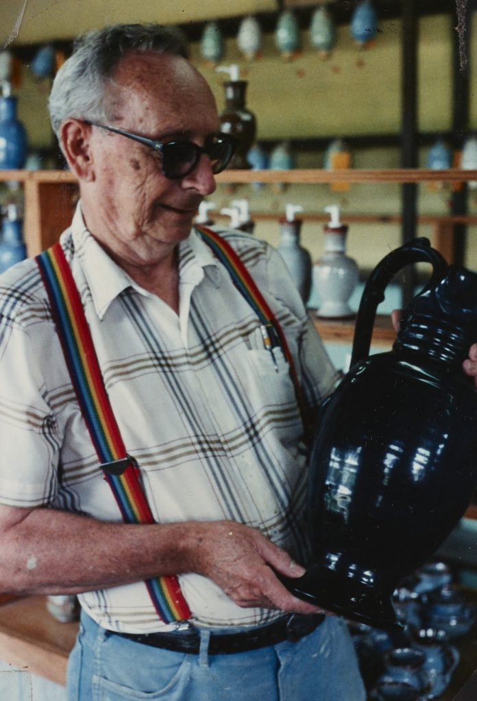 Older man identified as Foister Cole wearing glasses, jeans, a light plaid shirt, and rainbow suspenders holding a piece of pottery that is all black.