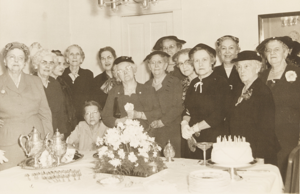 A group of Tea and Topics Book Club members at the twenty-fifth anniversary of the book club. The black-and-white photograph shows 14 group members standing around a table filled with a floral arrangement and cake.