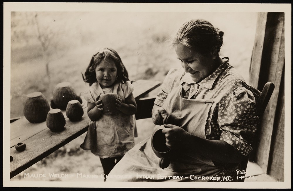 A child (left) holding pottery in their hands next to a table with more pottery on top. To the child's right is a woman identified as Maude Welch sitting in a rocking chair with an apron on, working on a pottery piece.