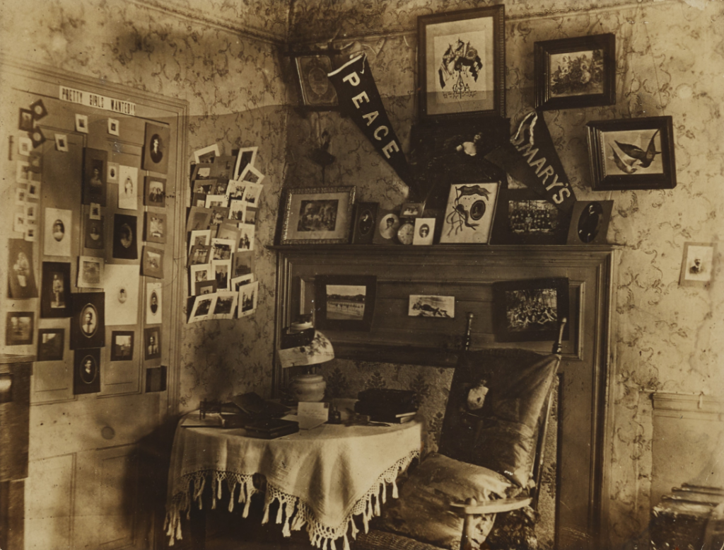 A dorm room at the University of North Carolina at Chapel Hill in 1903. The sitting area of the student's dorm room is decorated with pictures and flags across the walls and door. Along the top of the door a white horizontal sign reads "Pretty Girls Wanted!!"