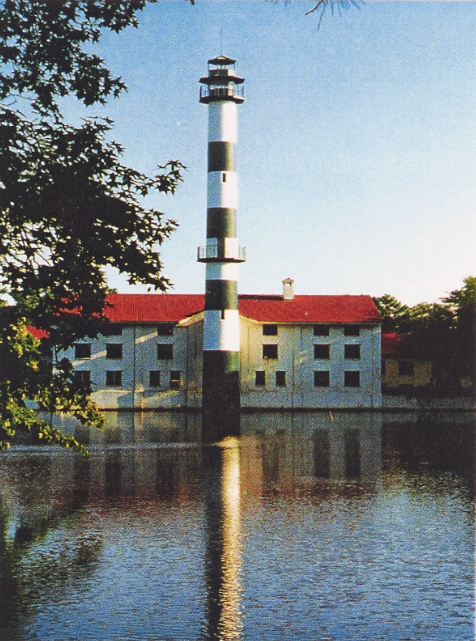 A picture of Mattamuskeet Lodge taken from across a body of water. A large white rectangular building sits on top of a body of water. The red roof of the structure is illuminated by sunlight. At the front of the building, a tall round tower extends from the water upwards. The green and white striped tower has outer decks towards its middle and top sections. Branches from a tree located in the lefthand side foreground partially obstruct the view of the lodge's left side.
