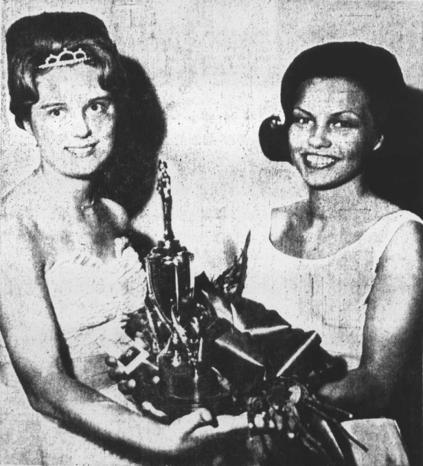 Two smiling teenagers stand side by side holding a trophy, ring, and flowers. On the left is Miss Ruth Nolen who is presenting the spoils of victory from the "Miss Ruby" contest to Miss Elaine Reece who is standing on the right.