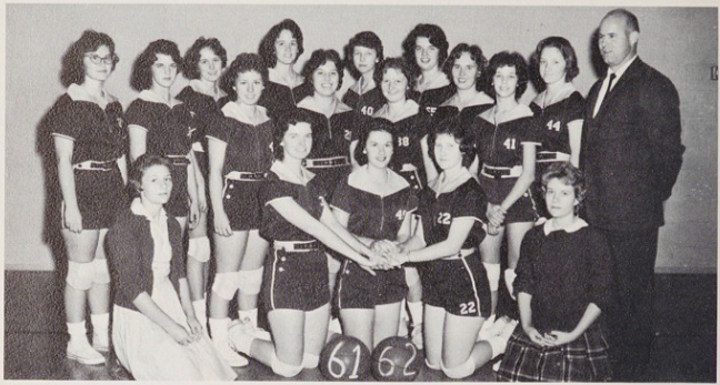 Individuals dressed in basketball uniforms both standing and kneeling for a picture. At the center on the floor are two basketballs that have the numbers 61 and 62 written on them. On the far right and bottom left are three individuals not dressed in basketball uniforms. The one standing on the far right is wearing a suit; bottom right is wearing a sweater and skirt; and bottom left is wearing either a top and skirt or a dress, along with a cardigan.