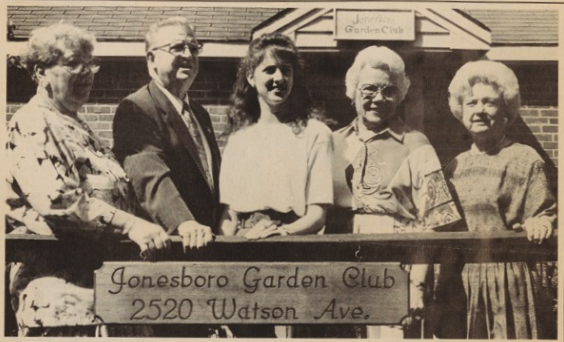 Five adults stand behind a wooden railing. There is a wooden sign attached to the railing that says, "Jonesboro Garden Club 2520 Watson Ave."