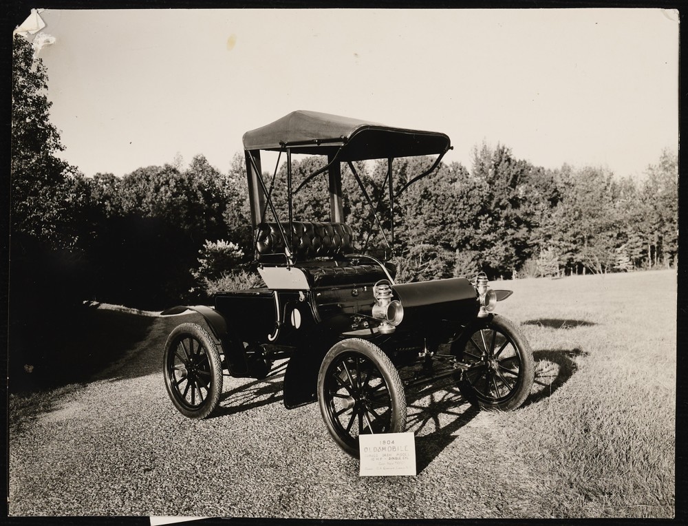 Image of a 1904 Oldsmobile with a sign propped against the front tire that reads: Curved dash model. 10 H. P. - single cyl. Cost new $650. Owner: O. A. Corriher. Landis, N.C.